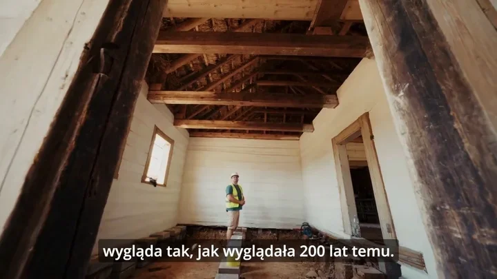 Pracownik w kamizelce ochronnej i kasku stoi we wnętrzu remontowanego drewnianego budynku z widocznymi belkami stropowymi.