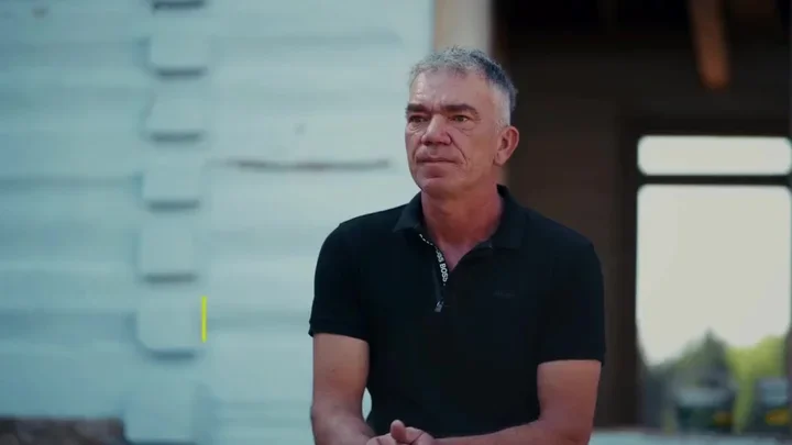 Mężczyzna w czarnej koszulce polo siedzi na zewnątrz przed drewnianą budowlą.