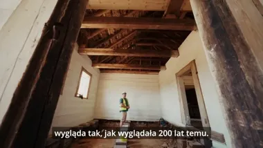 Pracownik w kamizelce ochronnej i kasku stoi we wnętrzu remontowanego drewnianego budynku z widocznymi belkami stropowymi.