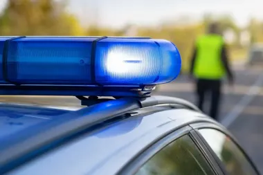 Błyskający niebieski kogut policyjny na dachu radiowozu, w tle rozmyta postać funkcjonariusza w kamizelce odblaskowej na drodze.